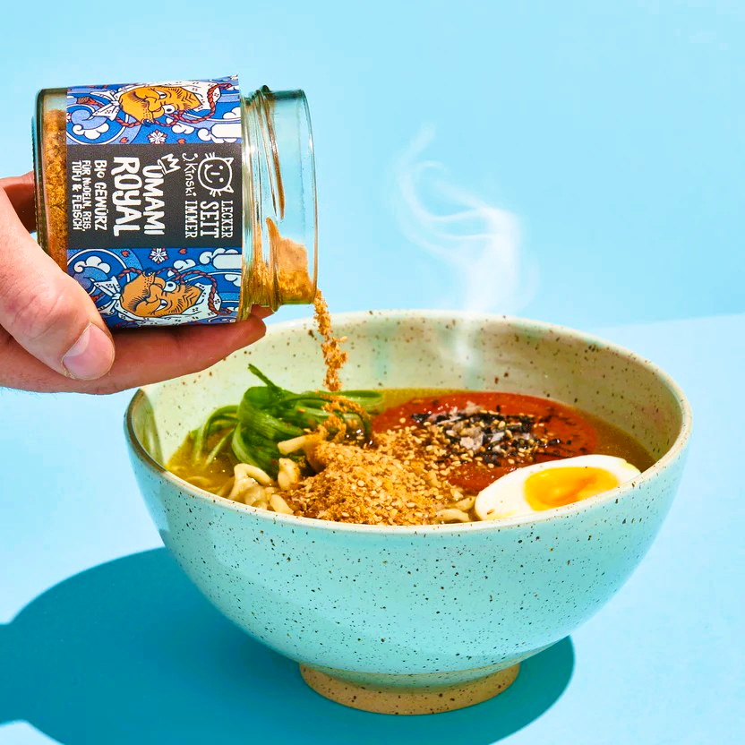 Schüssel mit Ramen: Eine große, flache Schüssel mit einer Portion Ramen-Nudeln.
Umami Royal Gewürz: Ein Glasgefäß mit dem Produkt "Umami Royal" wird über die Ramen gehalten.
Dampf: Aus der Schüssel steigt Dampf auf, was auf eine heiße Suppe hinweist.
Zutaten: In der Schüssel sind neben den Nudeln weitere Zutaten wie ein halbes gekochtes Ei, Gemüse und Toppings zu erkennen.