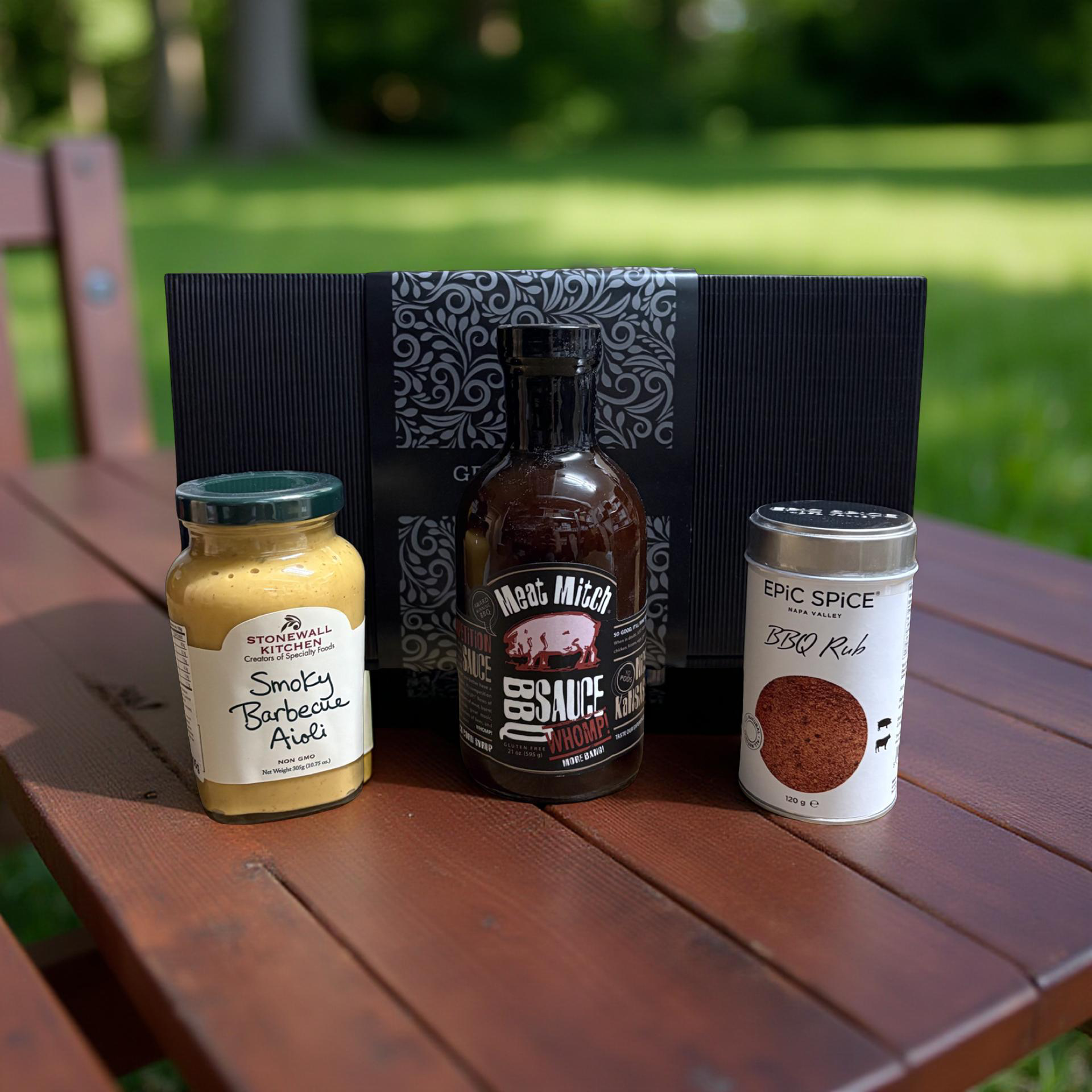 "Geschenkset für Fleischliebhaber mit Gourmet-BBQ-Saucen und Gewürzmischung in einer schwarzen Geschenkbox" mit Hintergrund im Freien auf einem braunen Holztisch