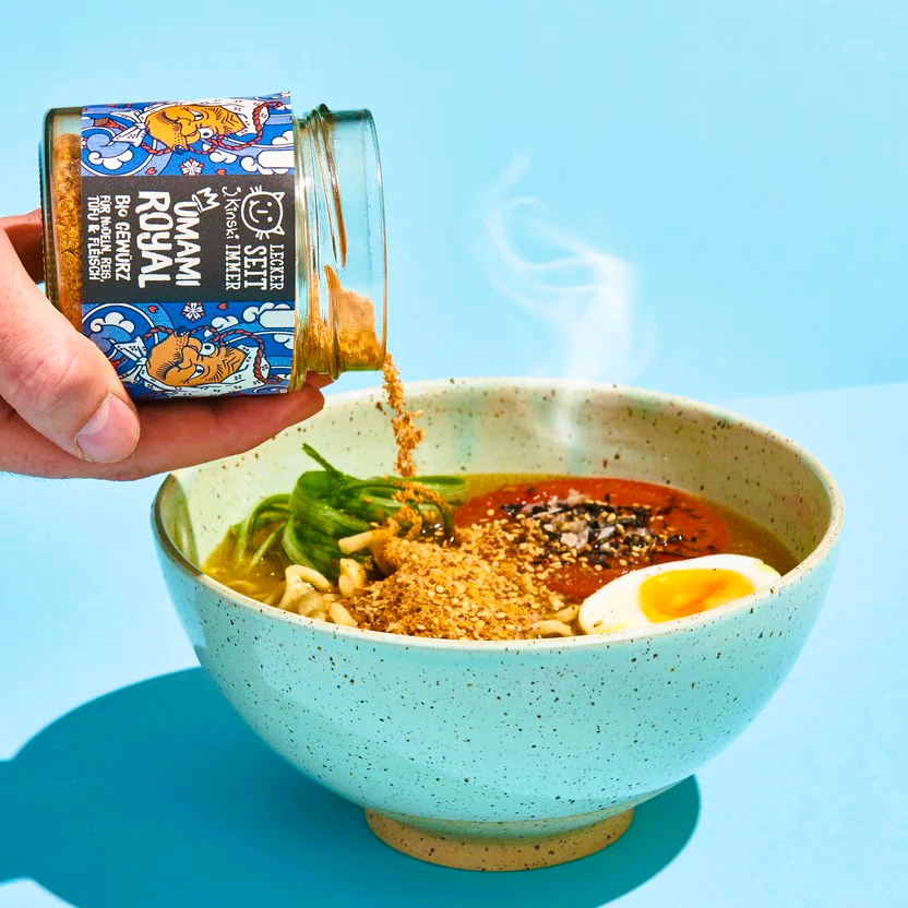 
                  
                    Schüssel mit Ramen: Eine große, flache Schüssel mit einer Portion Ramen-Nudeln.
Umami Royal Gewürz: Ein Glasgefäß mit dem Produkt "Umami Royal" wird über die Ramen gehalten.
Dampf: Aus der Schüssel steigt Dampf auf, was auf eine heiße Suppe hinweist.
Zutaten: In der Schüssel sind neben den Nudeln weitere Zutaten wie ein halbes gekochtes Ei, Gemüse und Toppings zu erkennen.
                  
                