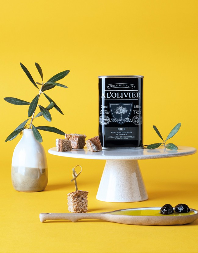 Olivenöl von A l'Olivier in schwarzer Metalldose auf gelbem Hintergrund mit Vase und OIlvenbaumzweige mit Aperohäppchen